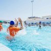 Persone in piscina giocano a pallanuoto, indossando caschi colorati.