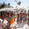 Festa in spiaggia con schiuma, adulti e bambini giocano sorridendo.