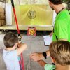 Bambino che pratica tiro con l'arco assistito da un adulto sotto una tenda.