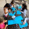 Bambini seduti a un tavolo all'aperto, colorando su fogli blu con pennarelli.