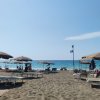 Spiaggia con ombrelloni beige e lettini, mare azzurro in lontananza. Pioggia di mare sullo sfondo.