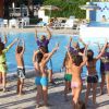 Persone partecipano a una lezione di esercizi accanto a una piscina. Tutti con le braccia sollevate.