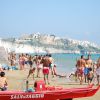 Spiaggia affollata con bagnanti e un pedalò rosso, vista su scogliera e città.