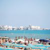 Spiaggia affollata con ombrelloni colorati, mare azzurro e un borgo sullo sfondo.