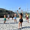 Persone giocano a beach volley su un campo sabbioso recintato, con colline sullo sfondo.