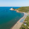 Spiaggia con file di ombrelloni azzurri, mare calmo e colline verdi.
