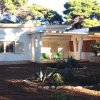 Una casa bianca con tetto sporgente, due sedie verdi sul portico e piante nel giardino.