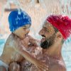 Uomo con cuffia rossa e bambino con cuffia blu giocano sotto una cascata in piscina.