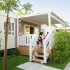 Veranda di una casetta prefabbricata con persone sedute sulle scale, circondata da alberi e arbusti.