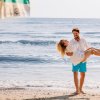 Un uomo solleva una donna sorridente su una spiaggia, davanti al mare calmo.