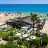 Vista aerea di una spiaggia con ombrelloni verdi e un lido con giardino e palme vicino al mare.