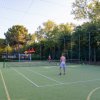Due persone giocano a tennis su un campo all'aperto, circondato da alberi e una rete di protezione.