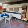 Una roulotte con una veranda arredata con tavolo, sedie bianche e un piccolo angolo cottura.