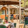 Persone sedute a tavoli all'aperto sotto tettoie di paglia, vicino a edifici arancioni e alberi.