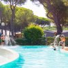 Bambini giocano in una piscina con giochi d'acqua e alberi intorno.