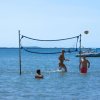 Persone giocano a pallavolo in acqua bassa con rete tesa. Barche sullo sfondo.