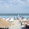 Spiaggia con ombrelloni azzurri e bianchi, mare sullo sfondo, cielo parzialmente nuvoloso.