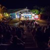 Pubblico seduto all'aperto guarda uno spettacolo serale su un palco.