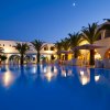 Piscina illuminata di notte, circondata da palme e edifici bianchi con archi, sotto un cielo sereno.