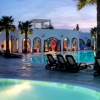 Piscina con arredo elegante e palme al tramonto, circondata da edifici bianchi con vetrate ad arco.