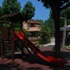 Parco giochi con scivolo rosso, altalene e giochi a molla. Alberi e recinzione sullo sfondo.