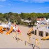 Parco giochi sulla spiaggia con scivoli colorati e altalene, circondato da alberi e cielo blu.