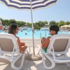 Due persone su sdraio sotto un ombrellone a bordo piscina, con giochi acquatici sullo sfondo.